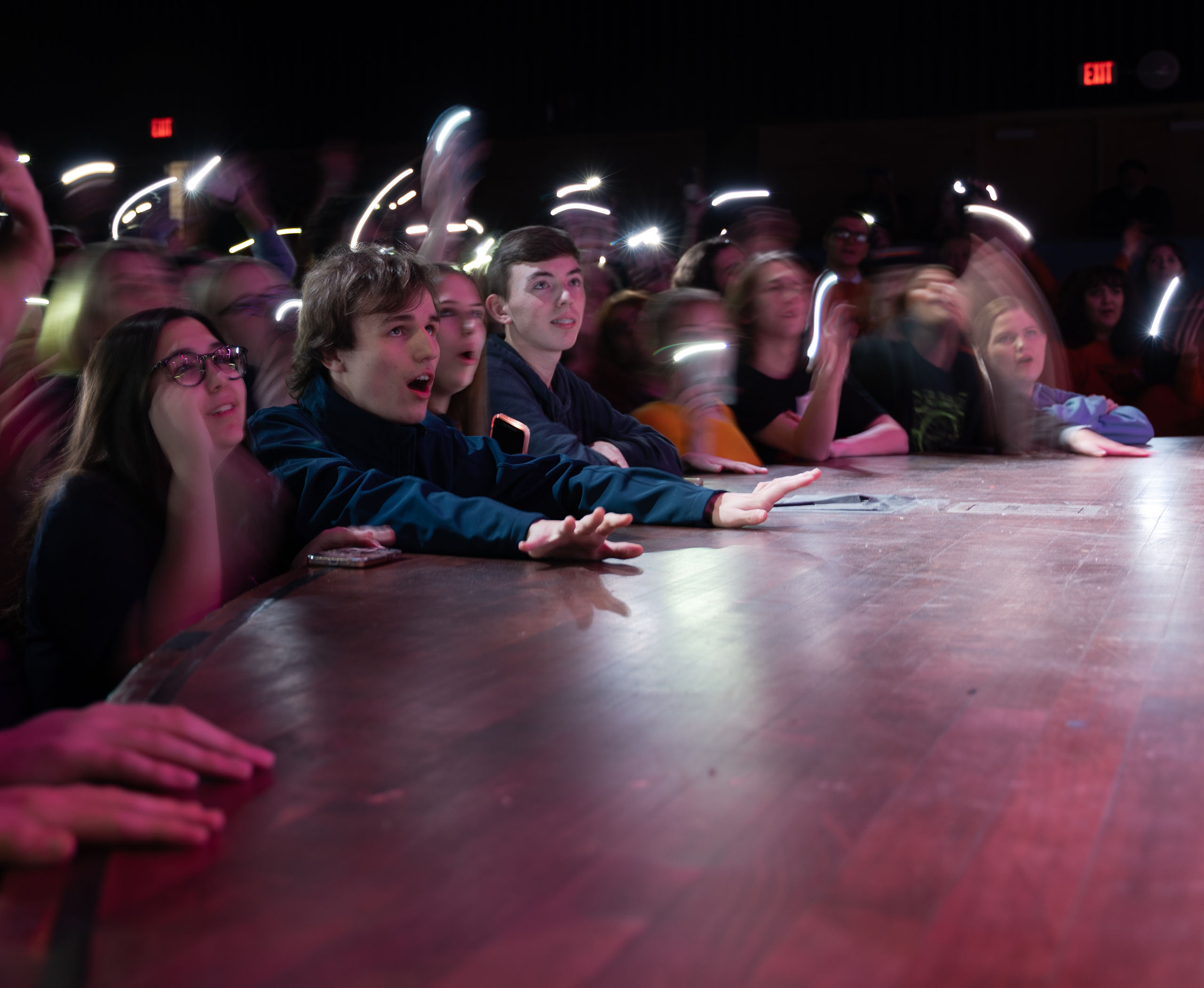 A group of students standing in the orchestra pit at the edge of a thrust stage. The students are waving phone flashlights and reaching onto the stage towards the performers.